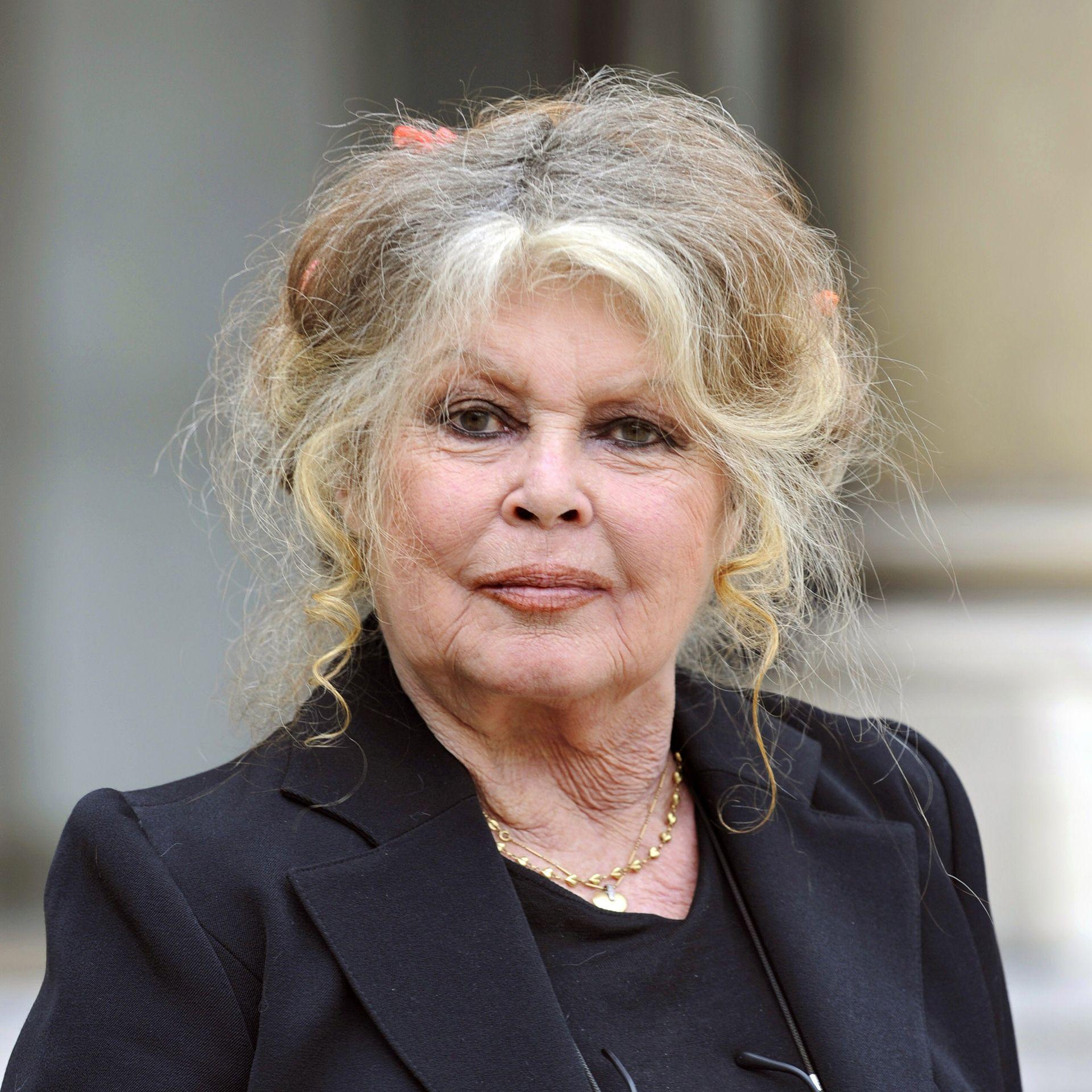 Brigitte Bardot fotografada em frente ao Pal&aacute;cio do Eliseu, vestindo um terno preto e com seus cabelos loiros levemente grisalhos.