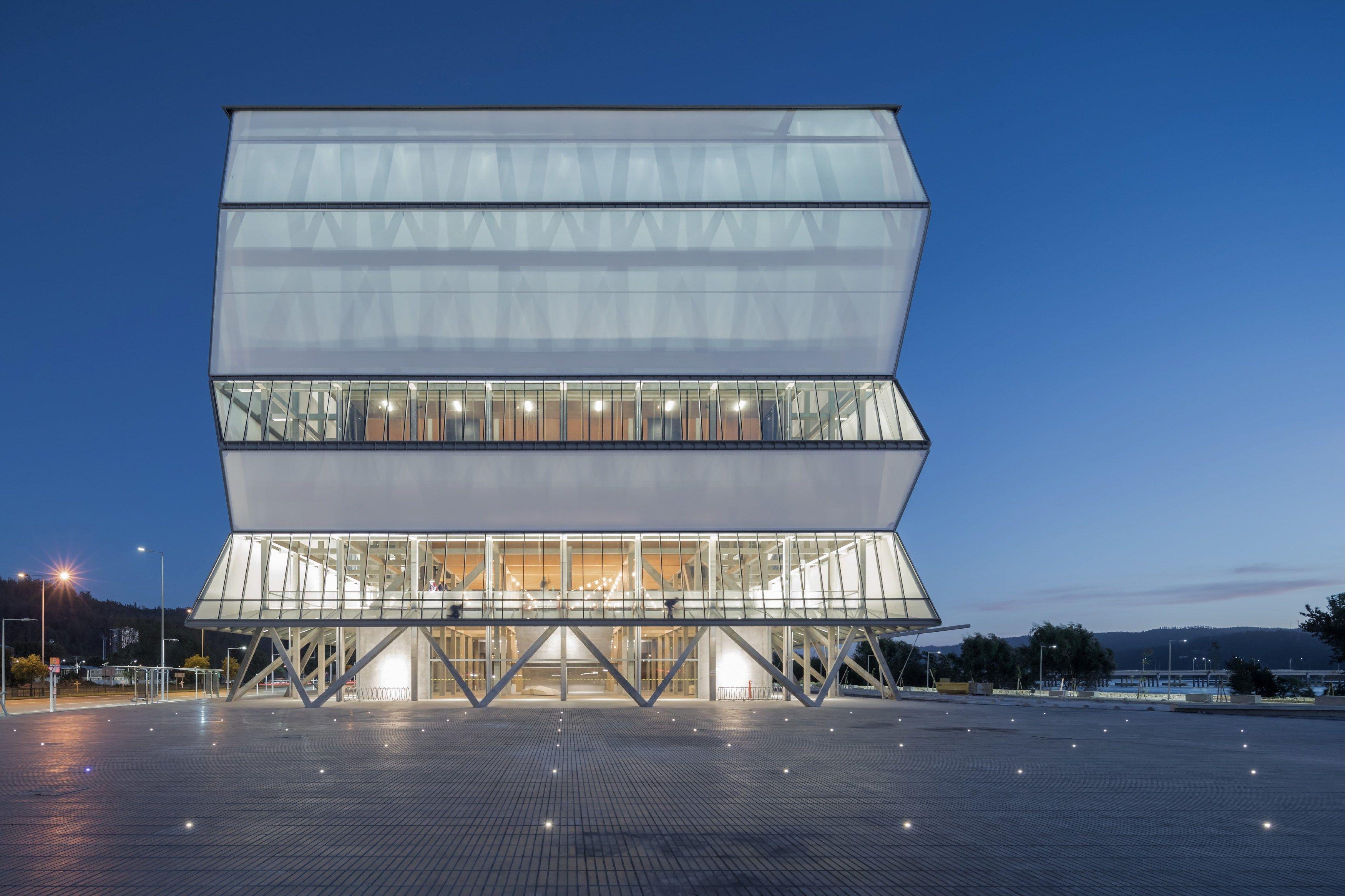 Edificio moderno iluminado al anochecer, con volumenes superpuestos y fachada translúcida frente a una amplia plaza pavimentada.