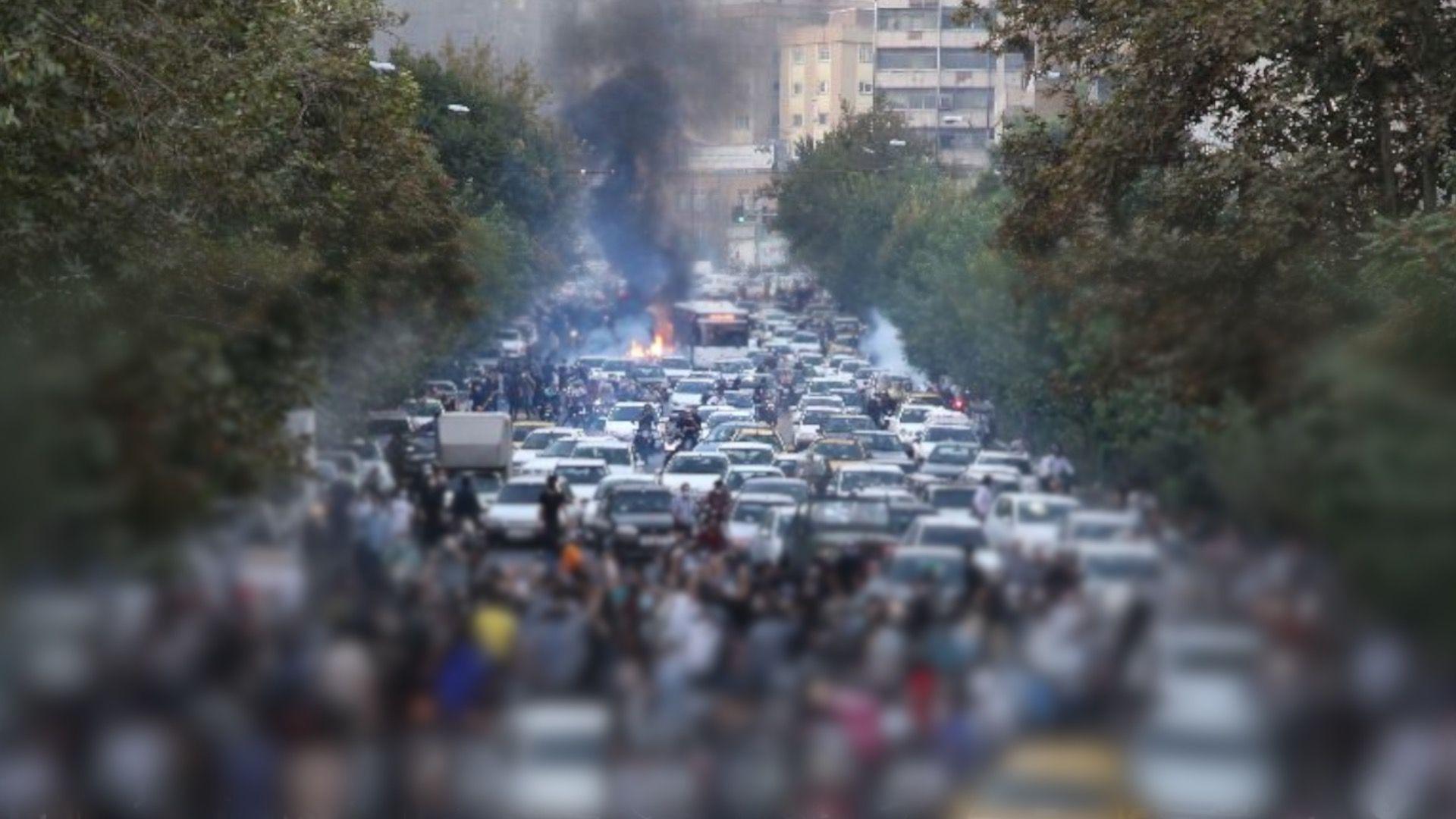 A wide street in Tehran blocked by vehicles, including cars and motorcycles, during the daytime on 21 September 2022. Dozens of people are gathered in front of and among the vehicles. A blur imposed by the BBC conceals the identities of people in the lower part of the image and nobody can be identified in the shot. A fire is burning in the background, with flames and dark black smoke billowing into the air. Green trees line both sides of the street. Tall buildings are in the background.