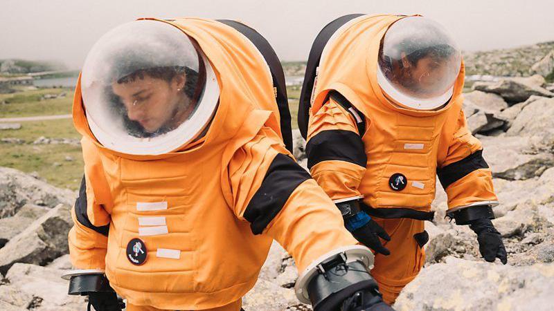 Duas pessoas de traje espacial laranja caminhando em meio às pedras