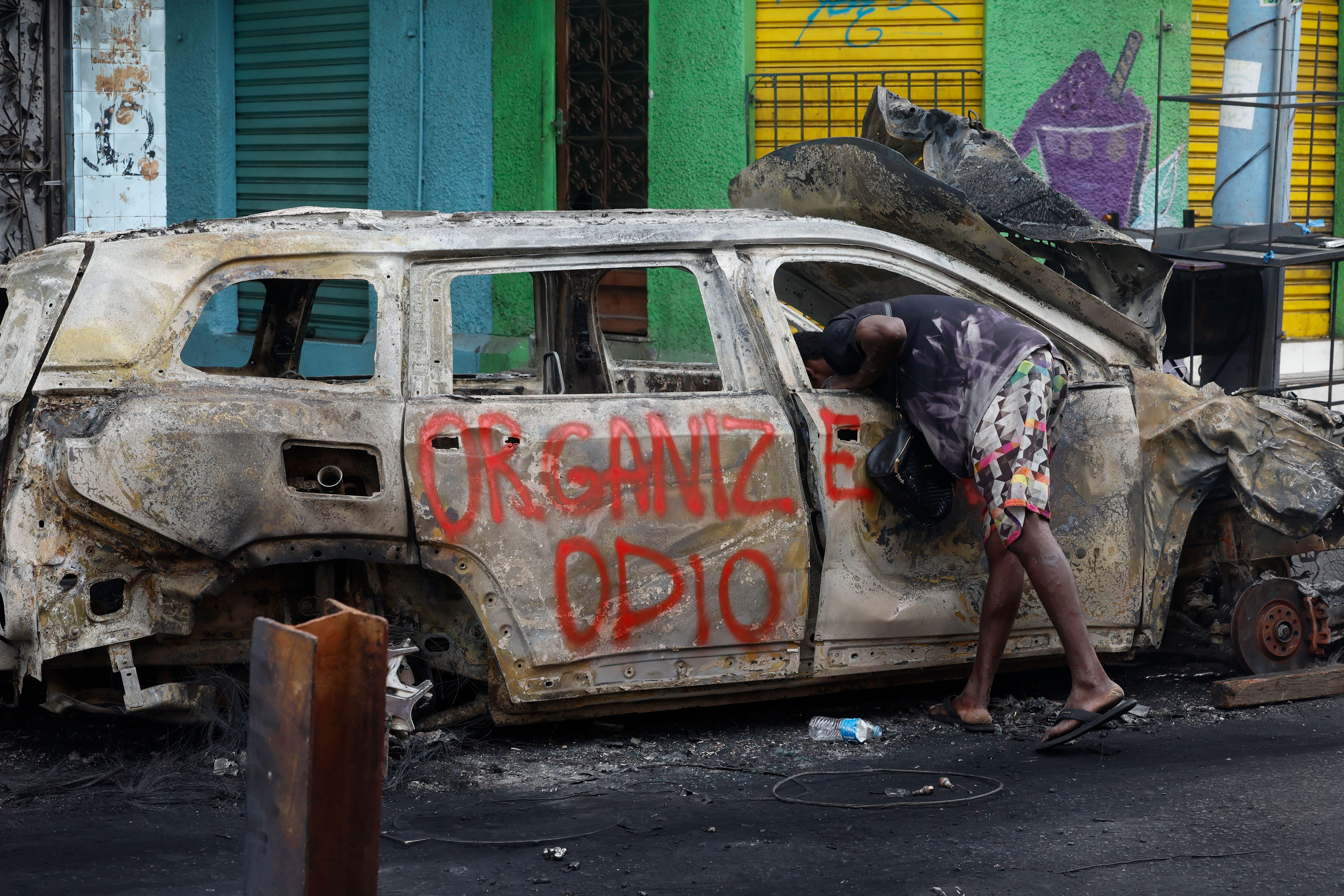 Un auto quemado con un grafiti que dice "organizar el odio" y una persona metiendo su cabeza por el hueco de la ventana de la puerta delantera izquierda.