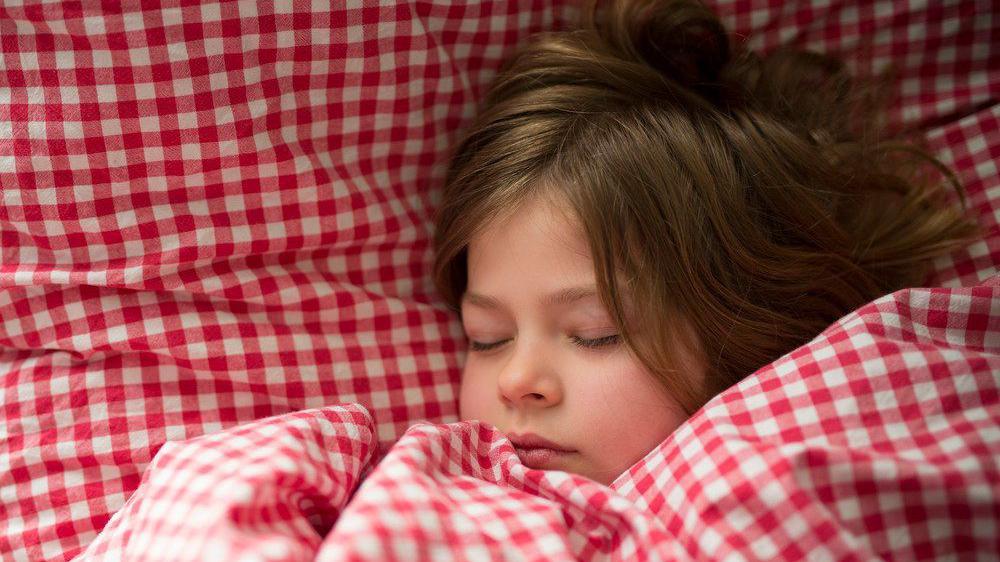 طفلة صغيرة تنام على سرير تحتها وسادة باللونين الأبيض والأحمر وتتلحف بغطاء باللونين الأحمر والأبيض.