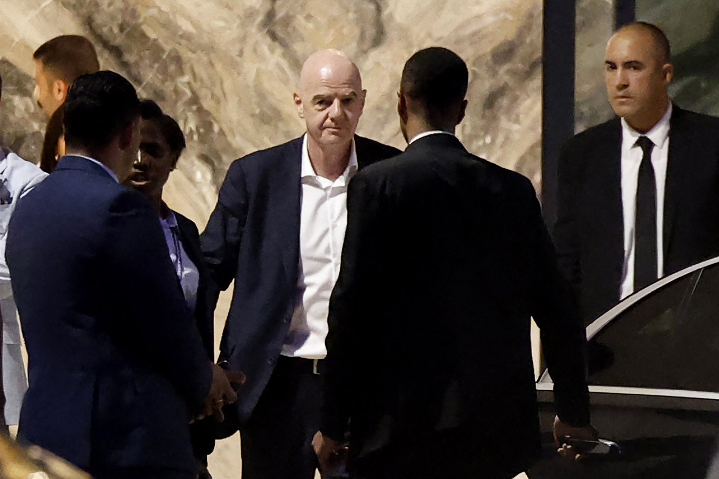 Infantino, en centro, está vestido con una camisa blanca y un traje oscuro. Está rodeado de varias personas. Un hombre tiene la puerta de un automóvil abierta

