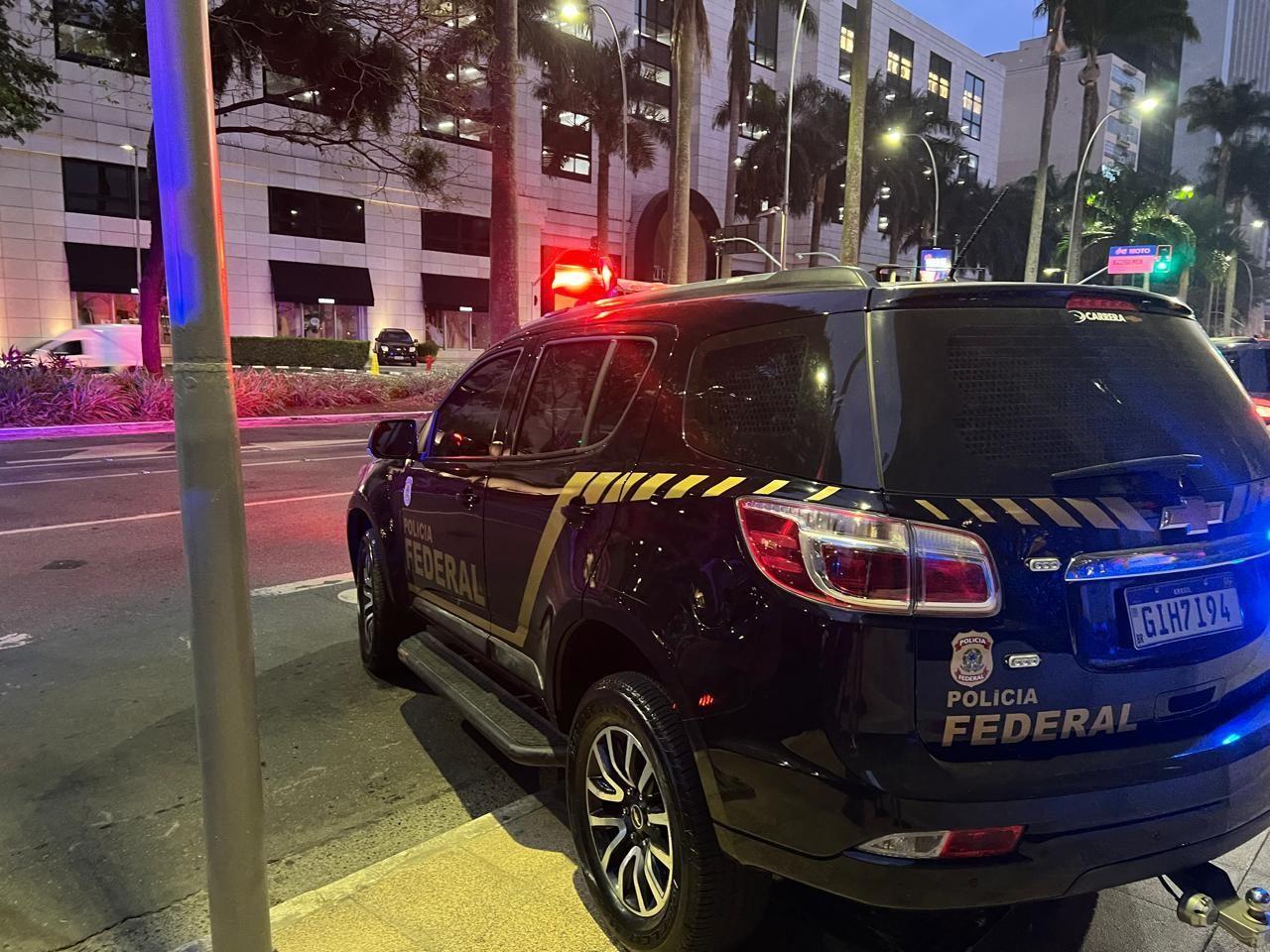 Foto de uma viatura preta com inscrição da Polícia Federal em frente a um prédio localizado em uma avenida ainda escura pela hora da madrugada
