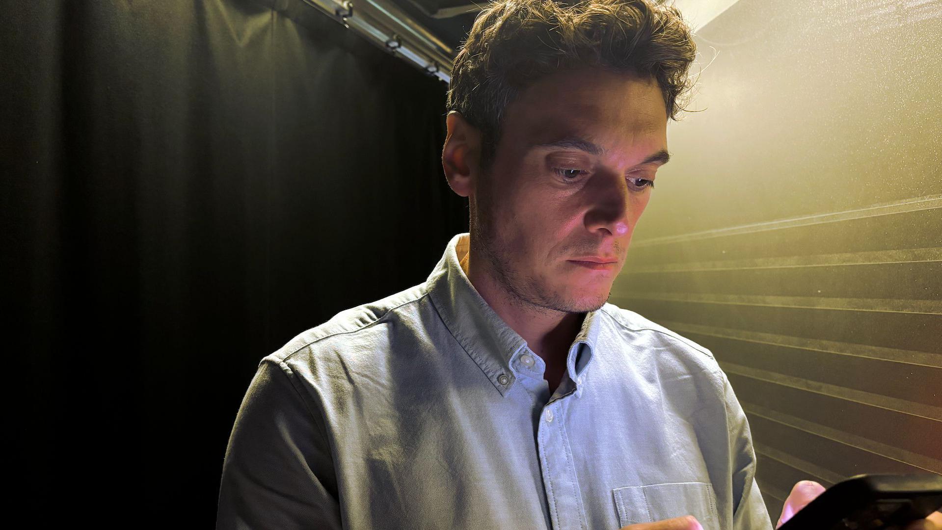Joe Tidy mirando un móvil. Tiene el pelo castaño corto y una camisa azul claro. 
