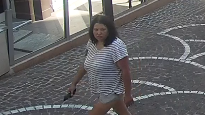 Una mujer de cabello oscuro y expresión seria mira hacia el otro lado de la calle por la que camina. Lleva una camisa a rayas blancas y negras. En una mano sostiene lo que parece ser un teléfono móvil.