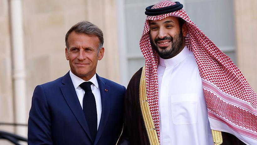 يظهر ماكرون مبتسماً، مرتدياً بدلة كحلية وربطة عنق سوداء، إلى جانب بن سلمان الذي يرتدي الزي السعودي التقليدي.