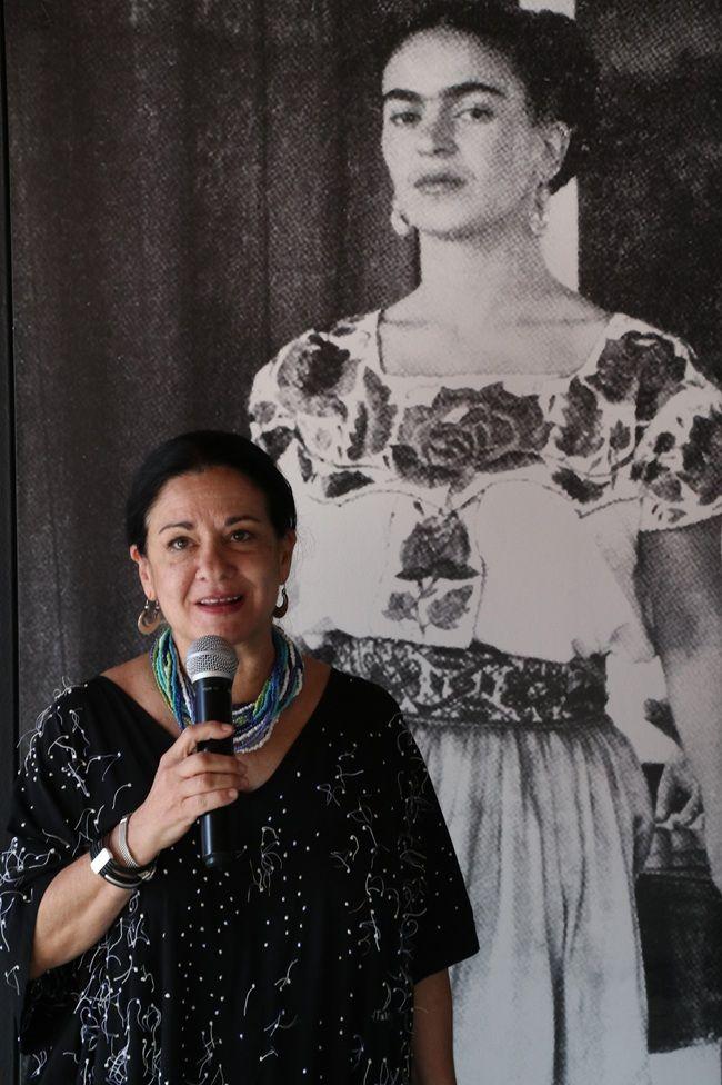 Cristina Kahlo Alcalá lleva un vestido negro con puntos blancos, aretes y un collar de varias vueltas azul y blanco. En su mano tiene un micrófono y detrás de ella hay una foto de Frida Kahlo, quien mira la cámara. Está vestida con la ropa tradicional de Tehuana
