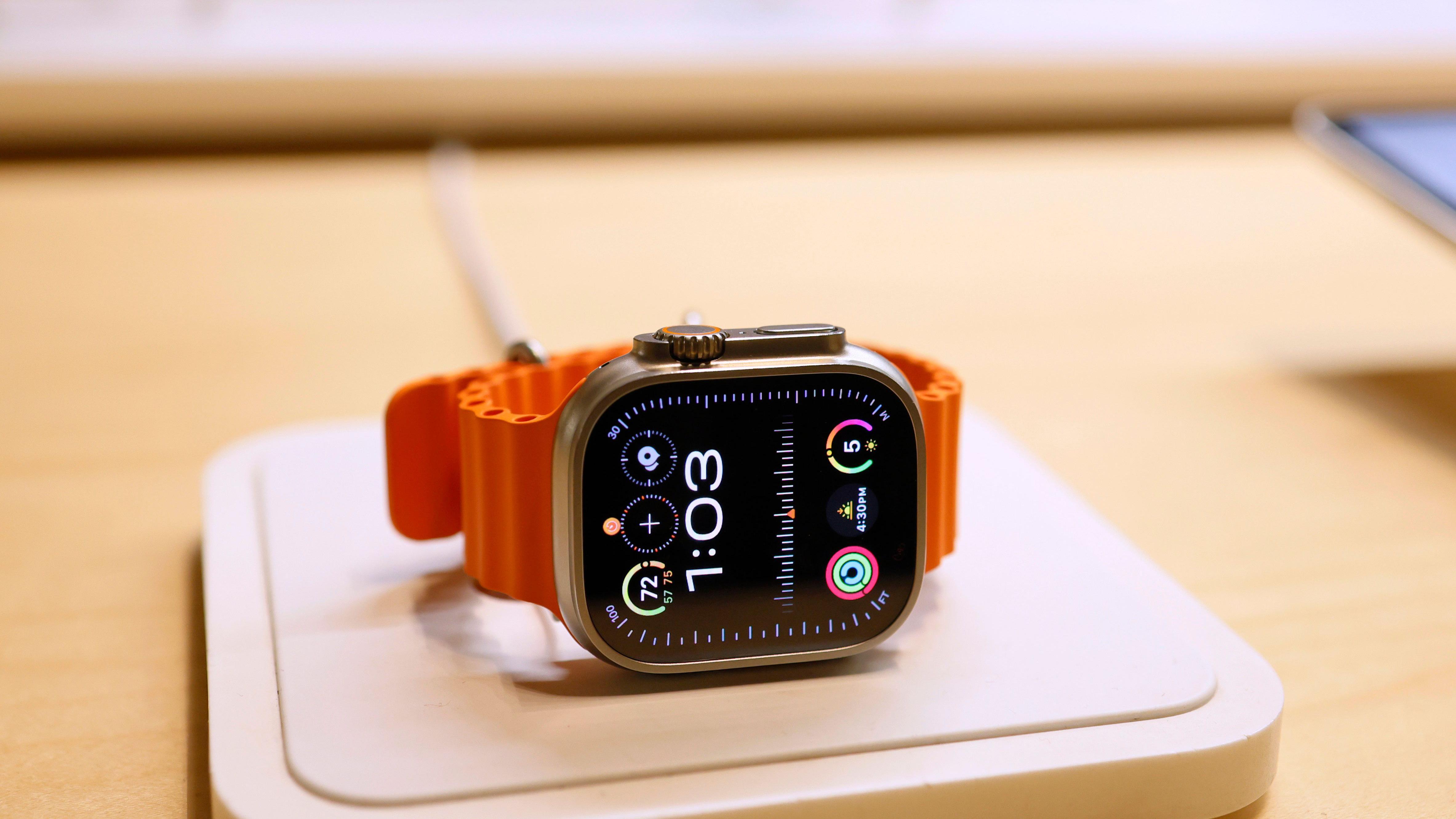 Apple Watch com mostrador preto e pulseira laranja sobre uma mesa, marcando 1h03