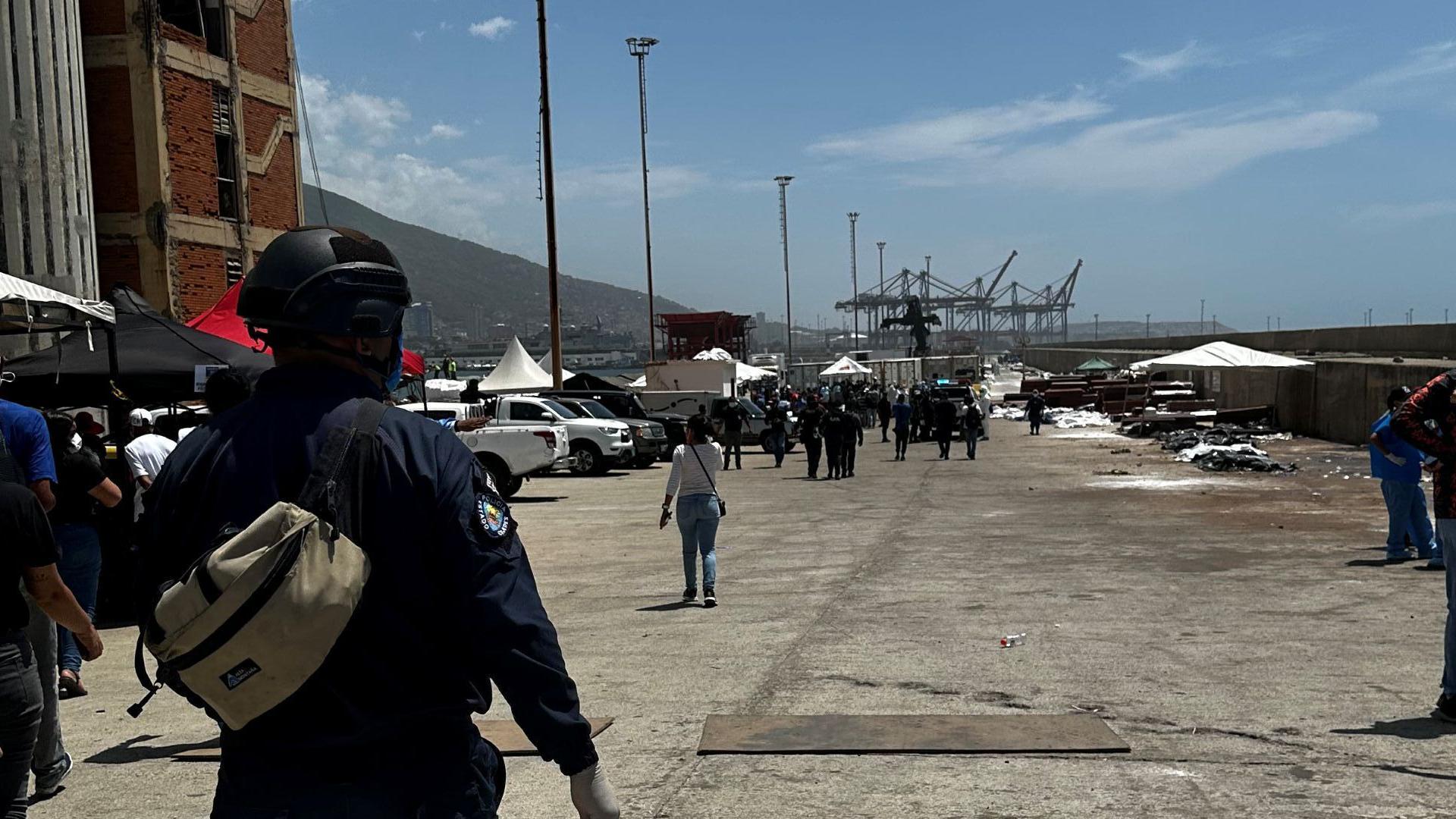 Un policía en primer plano de espaldas y al fondo se ven cuerpos tirados cubiertos con bolsas de plástico. 