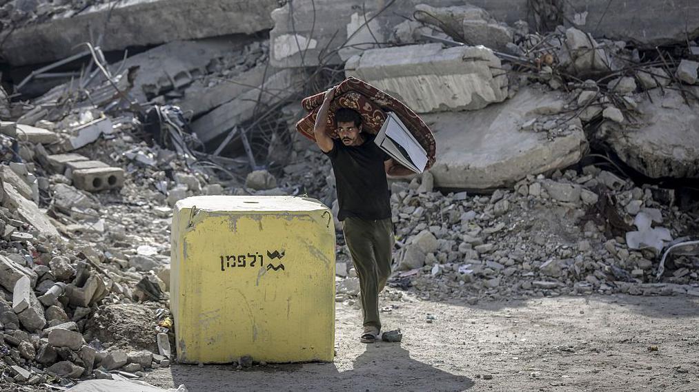 رجل يحمل مرتبة مطوية في غزة يمر بجوار كتلة خرسانية صفراء وضعها الجيش الإسرائيلي، وتظهر الأنقاض في الخلفية