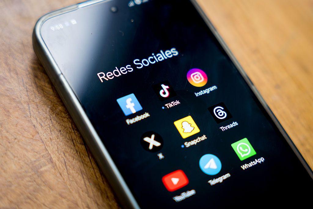 موبايل (هاتف محمول) لون الشاشة أسود وعليها مجموعة من تطبيقات التواصل الاجتماعي مثل واتس اب وتلغرام وإنستغرام... على طاولة خشبية.