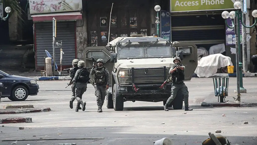 جنود إسرائيليون يطلقون الرصاص الحي والغاز المسيل للدموع من أسلحة وخلفهم مركبة عسكرية إسرائيلية.