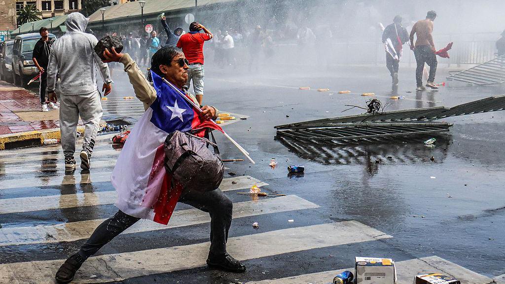 Protestas sociales de pescadores en Chile. Una persona cubierta con la bandera chilena lanza una piedra grande a la policía