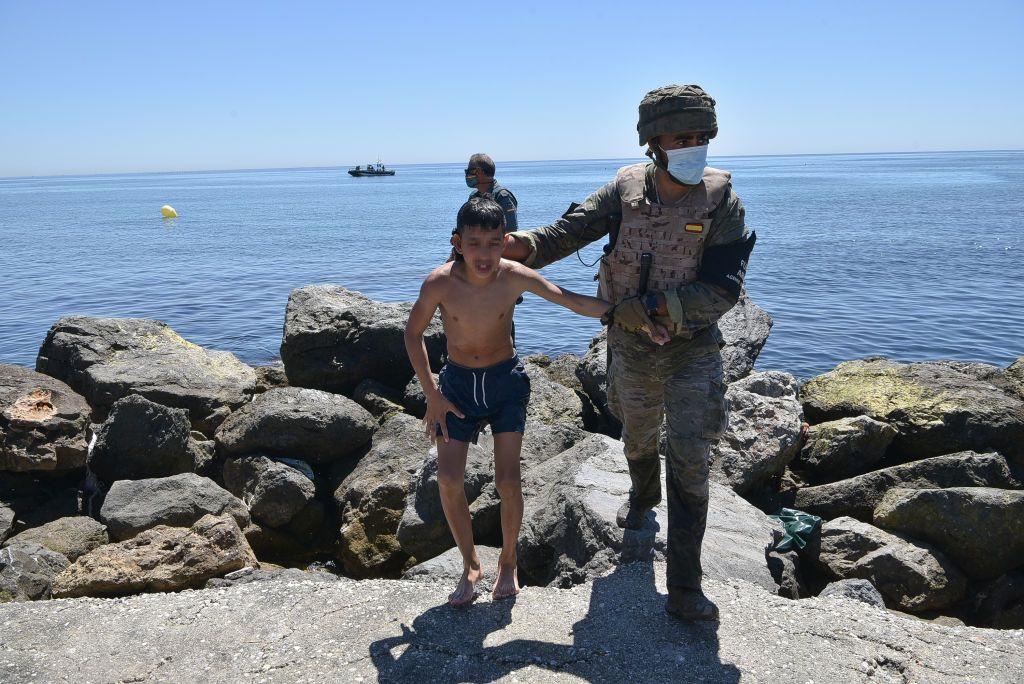 Un niño en traje de baño camina de la mano de un soldado español protegido con casco y mascarilla con el mar y una bahía rocosa a sus espaldas. Bajo el cielo soleado, en segundo plano se distingue a otro uniformado español. 
