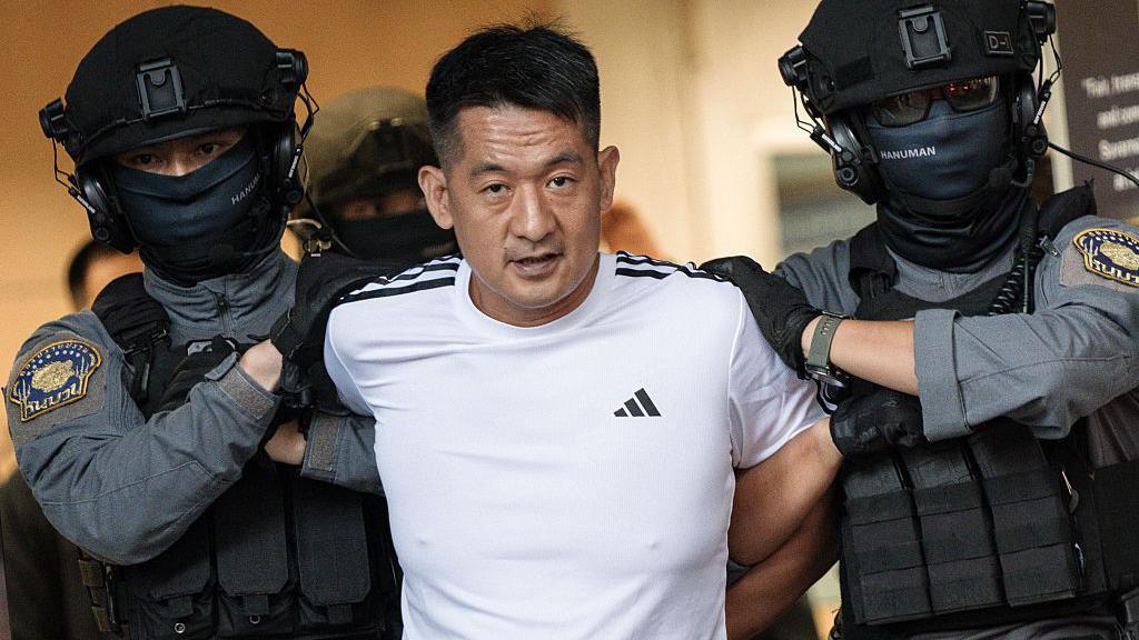 一名身穿白色阿迪達斯T 恤的男子，被兩名頭戴黑色頭盔、穿著防護背心的警衛押送