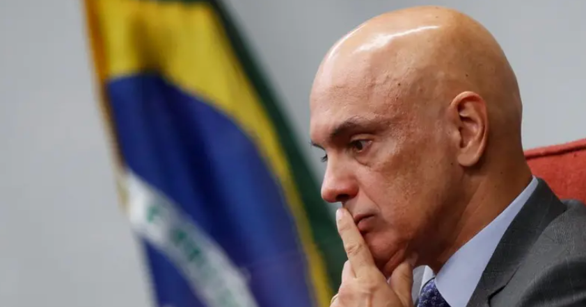 Foto de Alexandre de Moraes com a mão no queixo e a bandeira do Brasil ao fundo