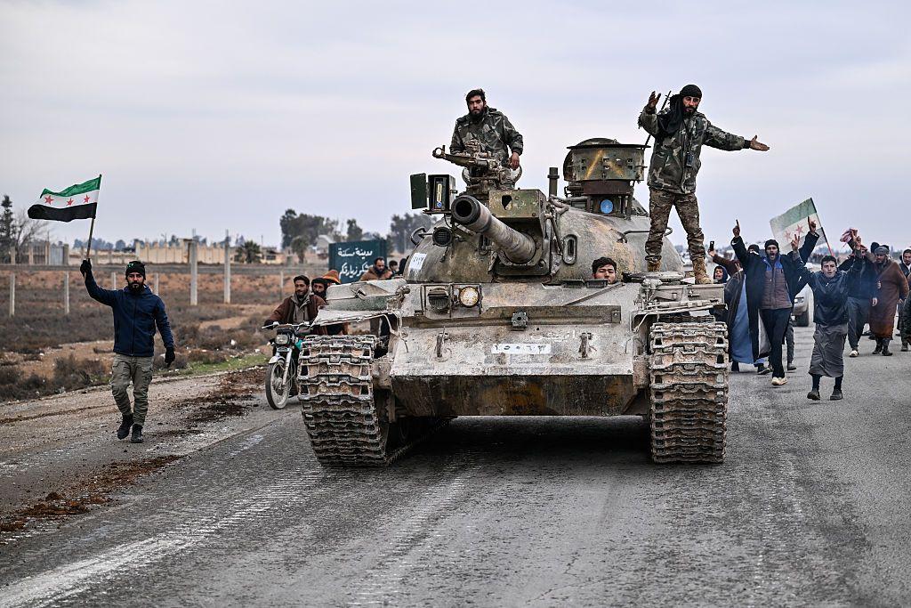 دبابة عليها عسكريون سوريون، ووراءهم عدد من الأشخاص يحملون علم سوريا ذو النجوم الثلاث الحمراء.