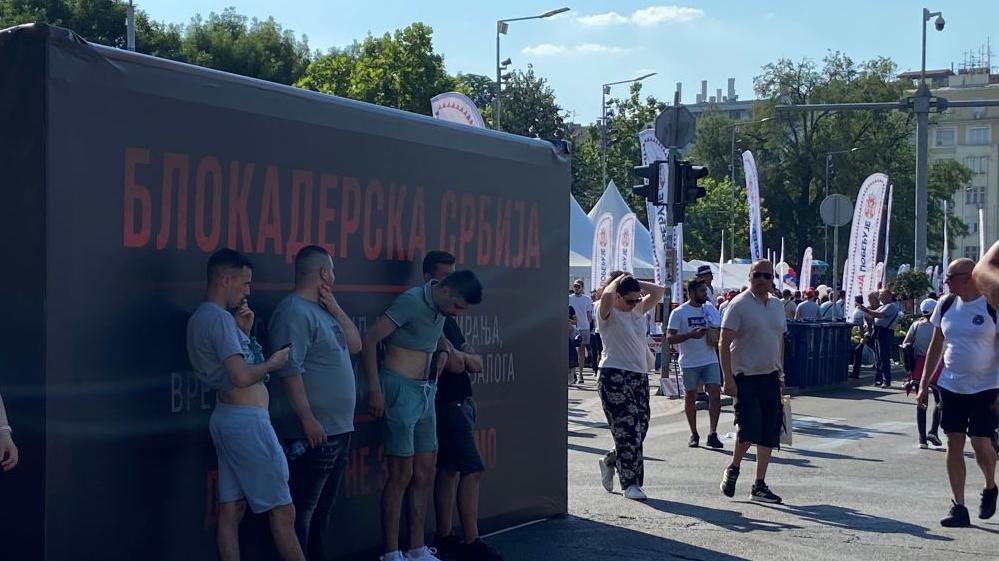 Ljudi na skupu vlasti stajali su u hladu pored improvizovane prostorije oblepljene natpisom 'Blokaderska Srbija'