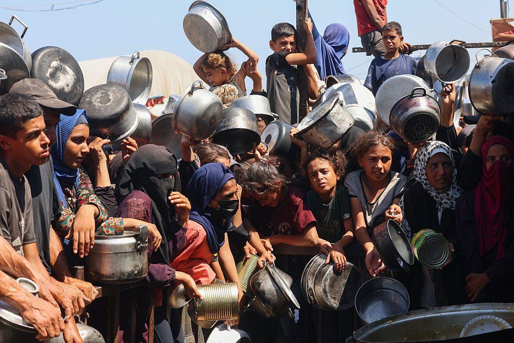 مجموعة من الأطفال والنساء يحملون أواني طعام فارغة ينتظرون على أمل الحصول على طعام. الصورة التقطت نهاراً.