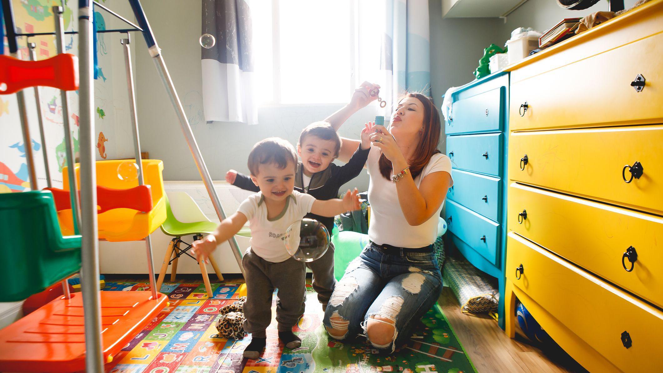 M&atilde;e brincando de bolinha de sab&atilde;o com os dois filhos. Ela est&aacute; ajoelhada no ch&atilde;o e os filhos sorriem e agitam os bra&ccedil;os, num quarto infantil bem colorido