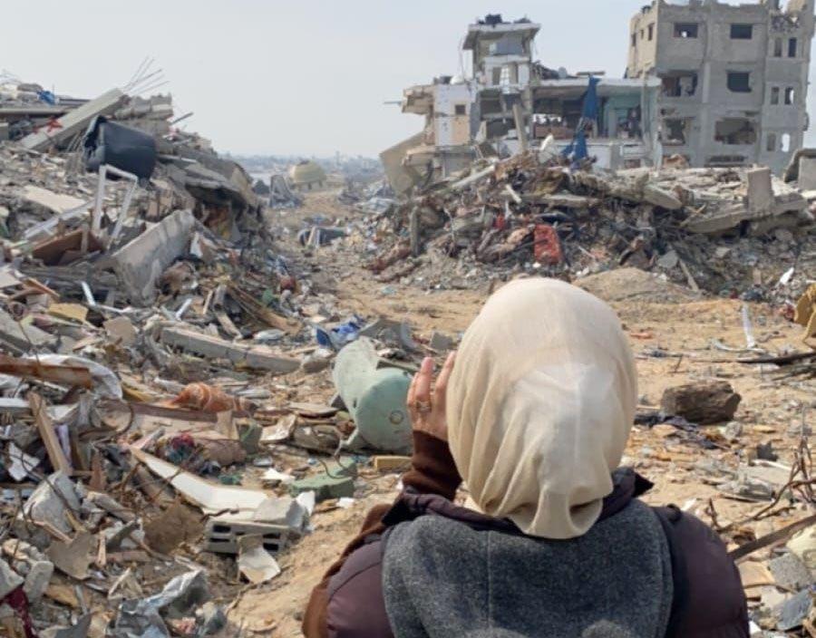Una mujer con un velo de color ligero y un abrigo marrón está de espaldas a la cámara. Frente a ella están las ruinas de dos edificios destruidos y grandes pilas de escombros y tierra   