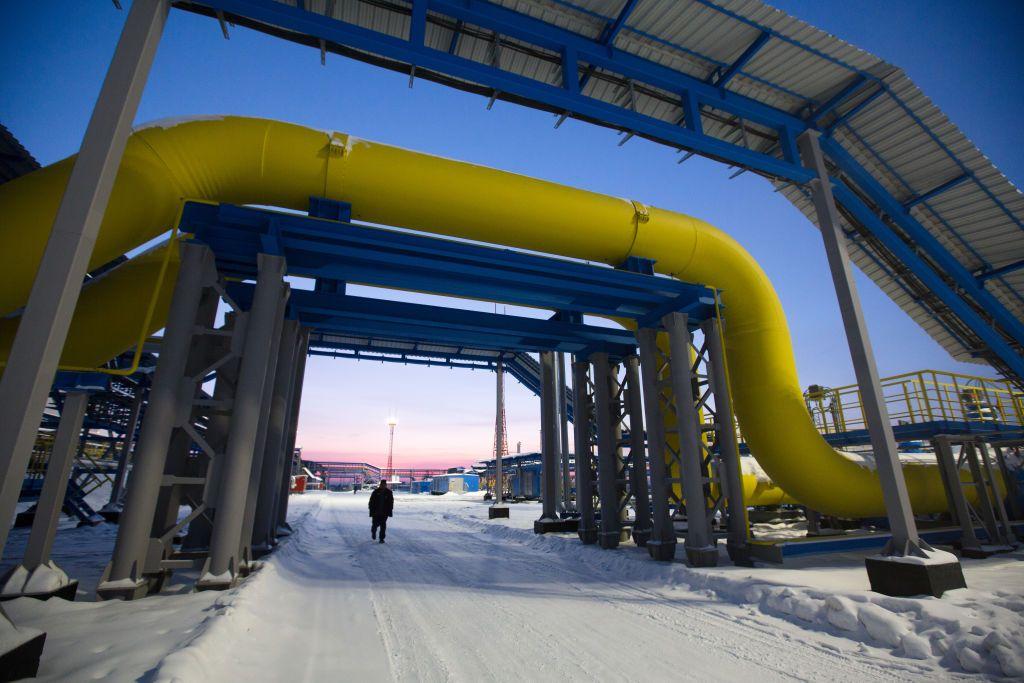 Infraestructura industrial con grandes tuberías amarillas elevadas sobre soportes metálicos en un entorno nevado, con una persona caminando debajo al atardecer.