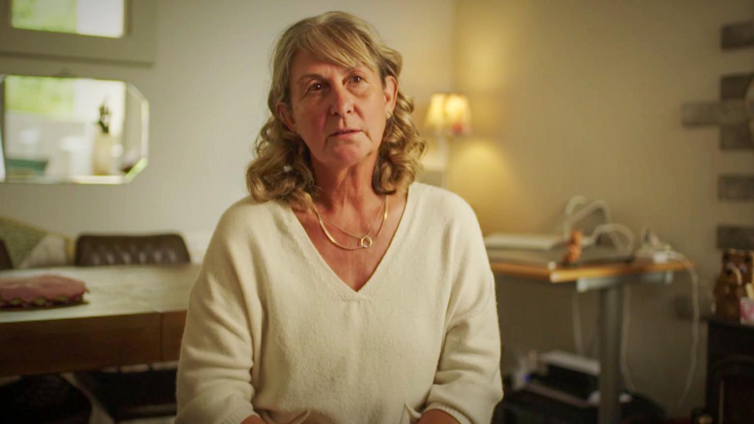 Photograph of Judith's neighbour, Michelle. She wears a white v neck jumper and has blonde shoulder length short hair. She wears two gold necklaces and looks sadly into the camera. A lamp can be seen illuminating the walls behind her. A dining table is behind her, as well as a desk behind her on the left. 