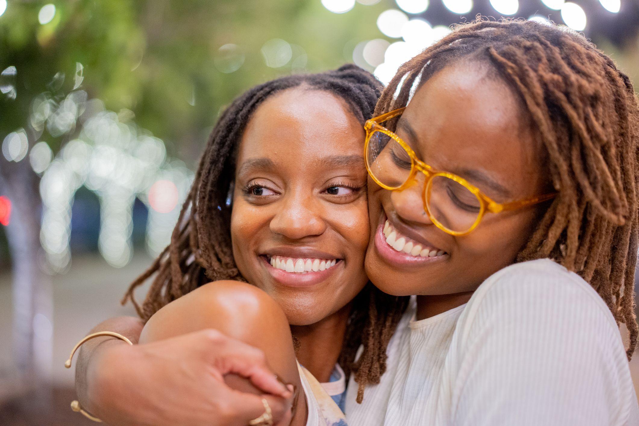 Duas irm&atilde;s g&ecirc;meas afro-americanas sorrindo, na casa dos vinte anos, abra&ccedil;ando-se carinhosamente 