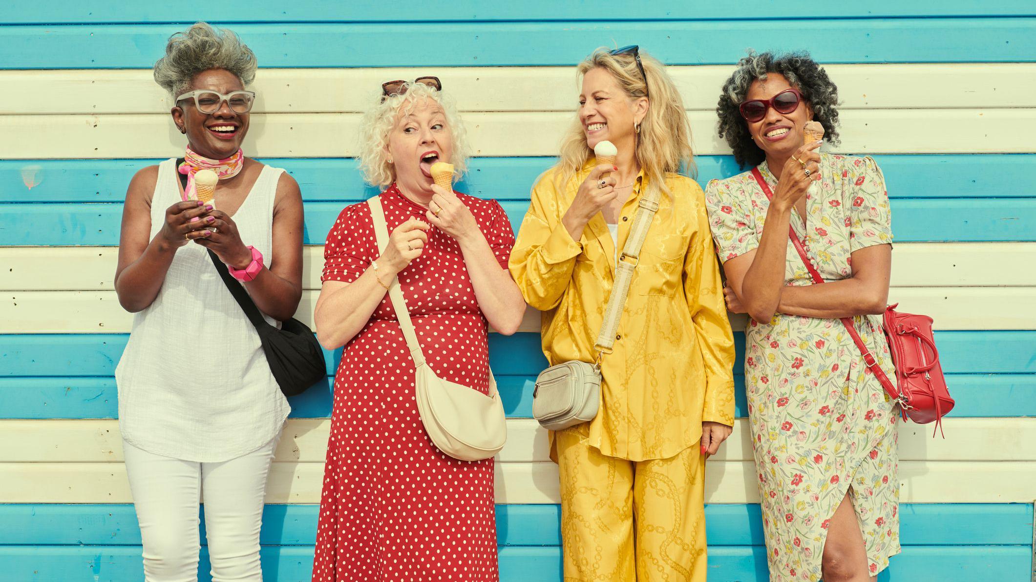 cuatro mujeres de pie frente a una pared de madera de color amarillo y azul. Todas sostienen un helado en sus manos y sonríen. 