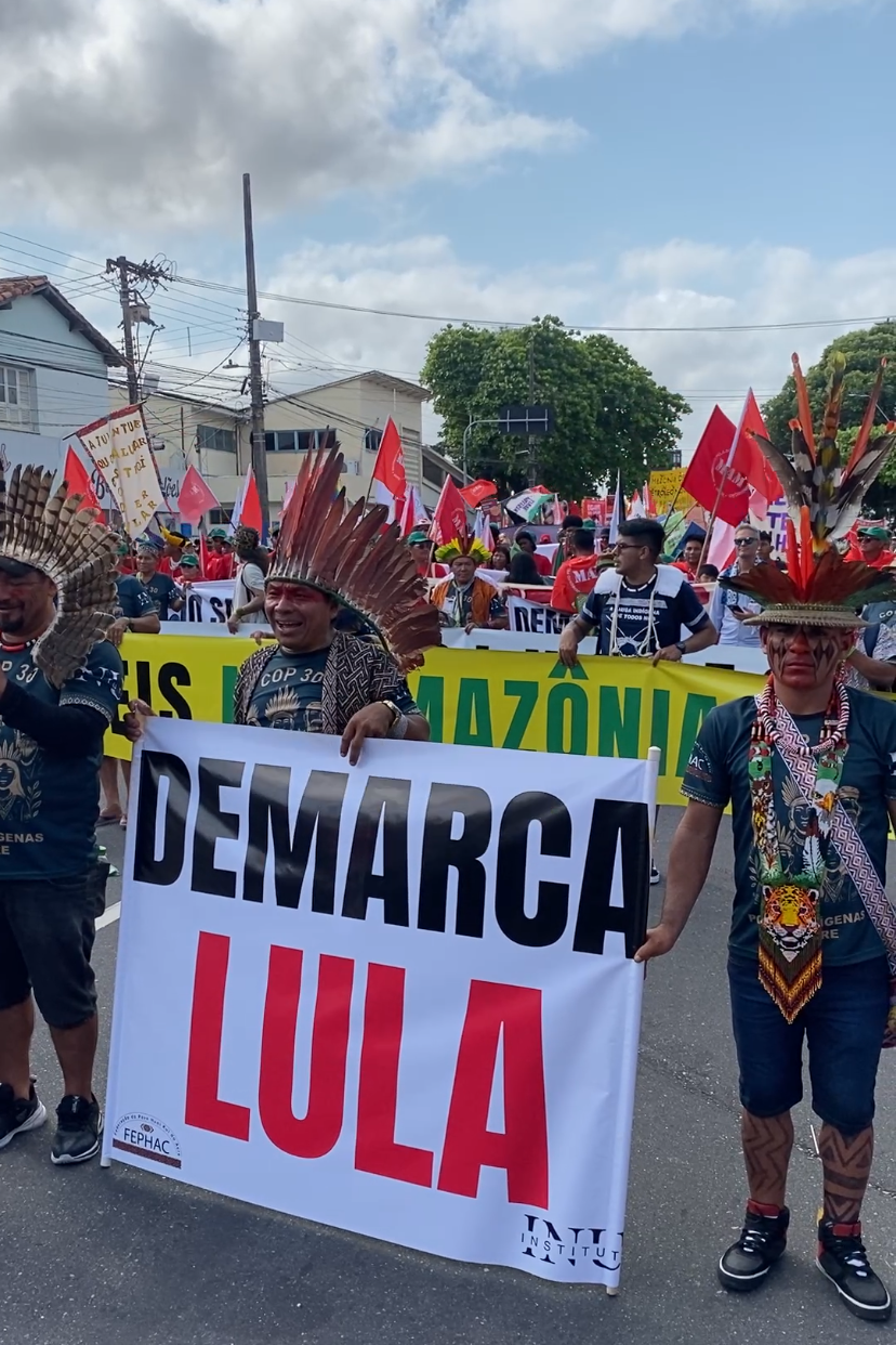 Manifestantes indígenas vestindo adereços tradicionais e segurando um cartaz onde se lê 