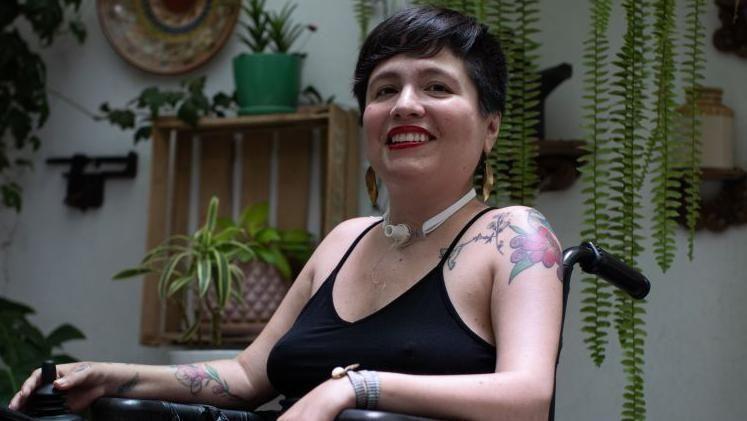 Ana, en primer plano, sentada en una silla de ruedas, con un hueco de traqueotomía en la garganta. De fondo hay una pared blanca con muchas plantas. 