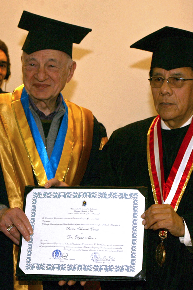 Edgar Morin con la toga y birrete y una medalla de cinta azul y amarilla sostiene el título junto al rector Juan Tutuy Aspauza, quien también llevaba toga y birrete y una medalla con cinta roja y blanca. 
