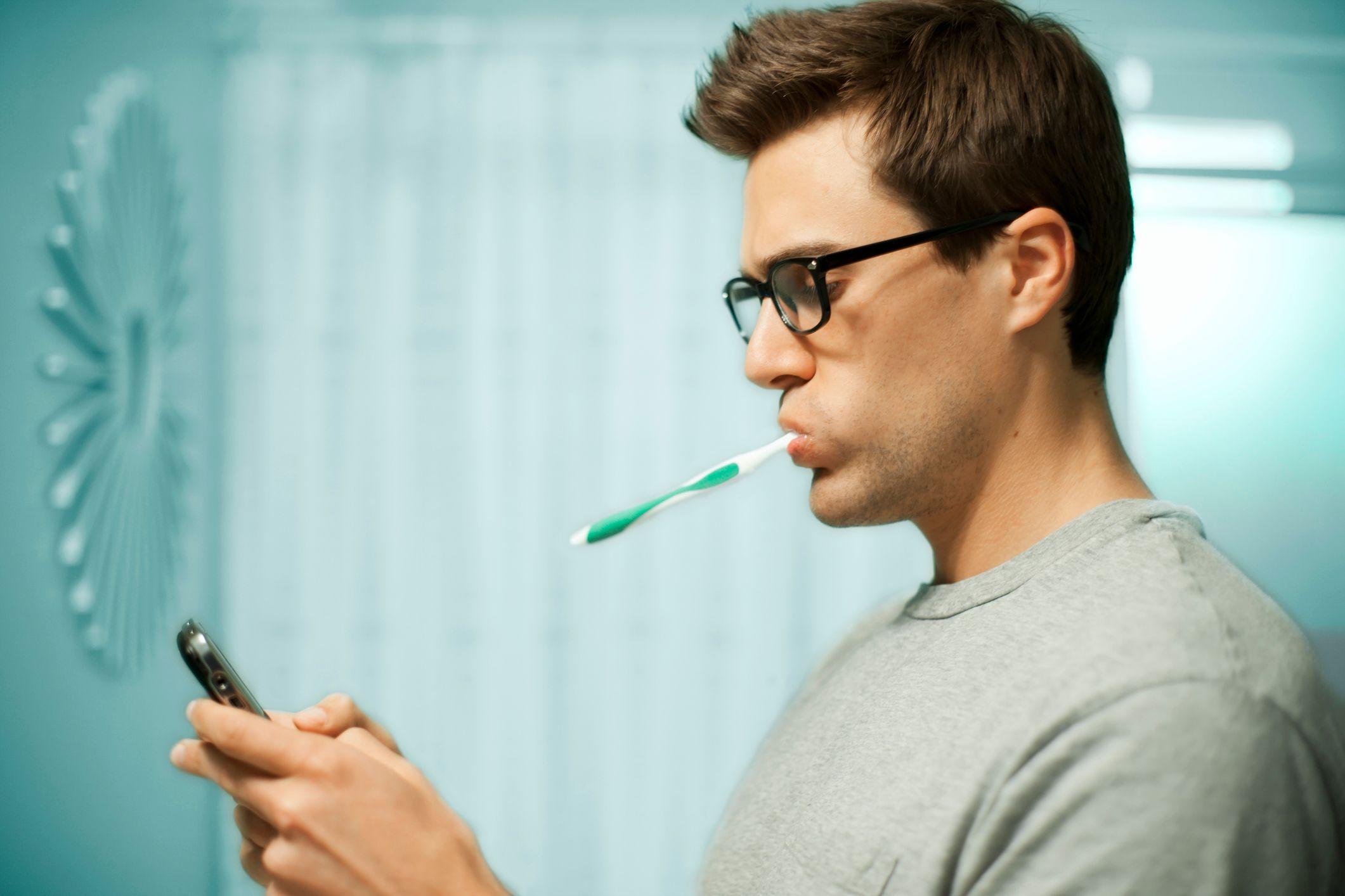 Un hombre de perfil en un baño, con anteojos y un cepillo de dientes en la boca, consulta su teléfono móvil