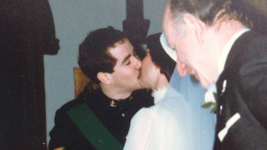 H&aacute; quatro pessoas nesta fotografia. No centro, um casal de noivos se beija no dia do casamento. O noivo veste um uniforme militar preto. A noiva, de costas para a c&acirc;mera, usa um vestido branco de gola alta e um v&eacute;u branco preso a um adorno de cabe&ccedil;a. Ambos t&ecirc;m cabelos escuros. &Agrave; esquerda do noivo est&aacute; o c&ocirc;nego, que celebrou o casamento. &Agrave; direita da noiva, um convidado do casamento veste um terno preto.