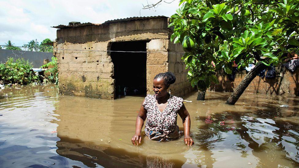 Uma mulher em p&eacute; com &aacute;gua at&eacute; a cintura, com um pr&eacute;dio inundado e uma &aacute;rvore atr&aacute;s dela.