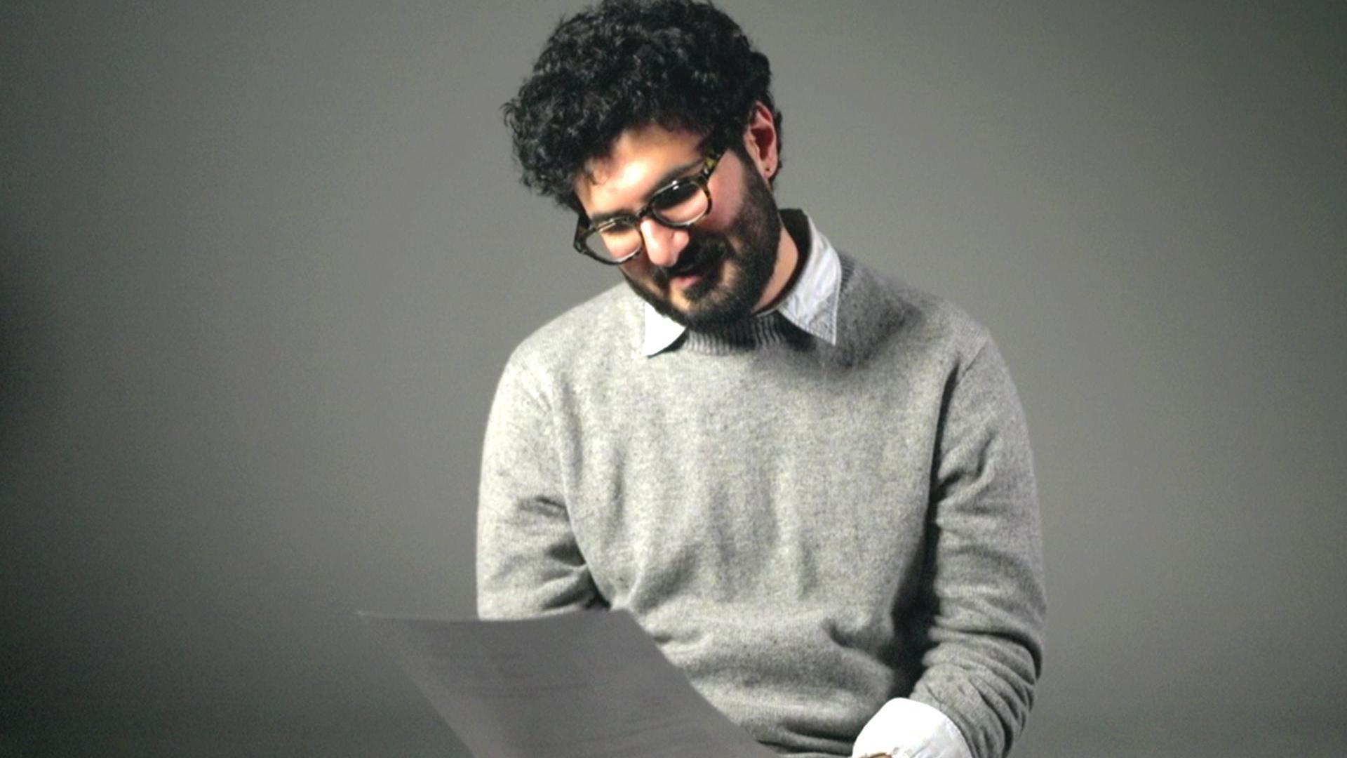 一個有著濃密捲曲的黑髮、鬍鬚和絡腮鬍的男人正在看著一張紙。他戴著眼鏡，穿著淺灰色套頭衫，內搭淺色襯衫，下身是淺棕色長褲。