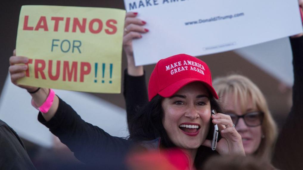 Una mujer con una gorra de Hagamos a Estados Unidos Grande Otra Vez sostiene un cartel que dice LATINOS POR TRUMP