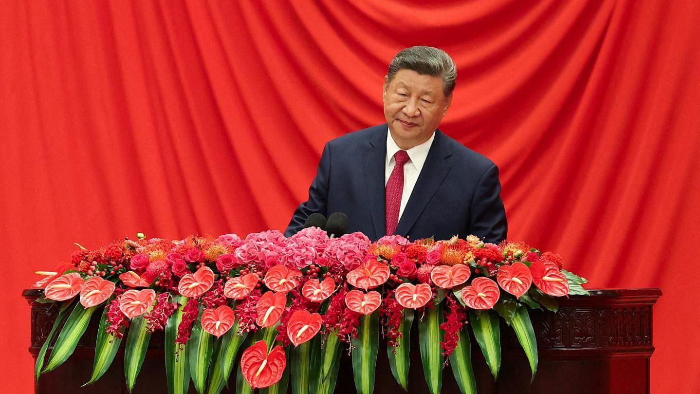 Xi Jinping, vestido con un traje oscuro, se encuentra frente a una cortina roja y detrás de un atril de madera cubierto de flores rojas y rosas brillantes y hojas verdes.
