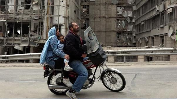 Una pareja viajando en motocicleta con un niño en medio. Detrás hay esdificios destruidos.