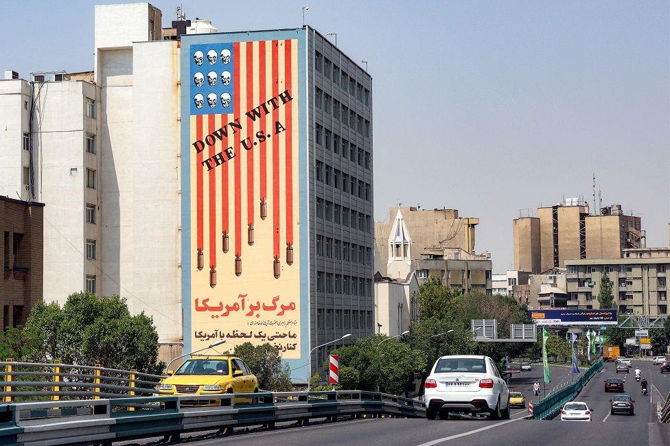 Vehículos transitan por una avenida que tiene a un lado un edificio con un mural que tiene la bandera de EE.UU. y un mensaje que dice "Down with the U.S.A"