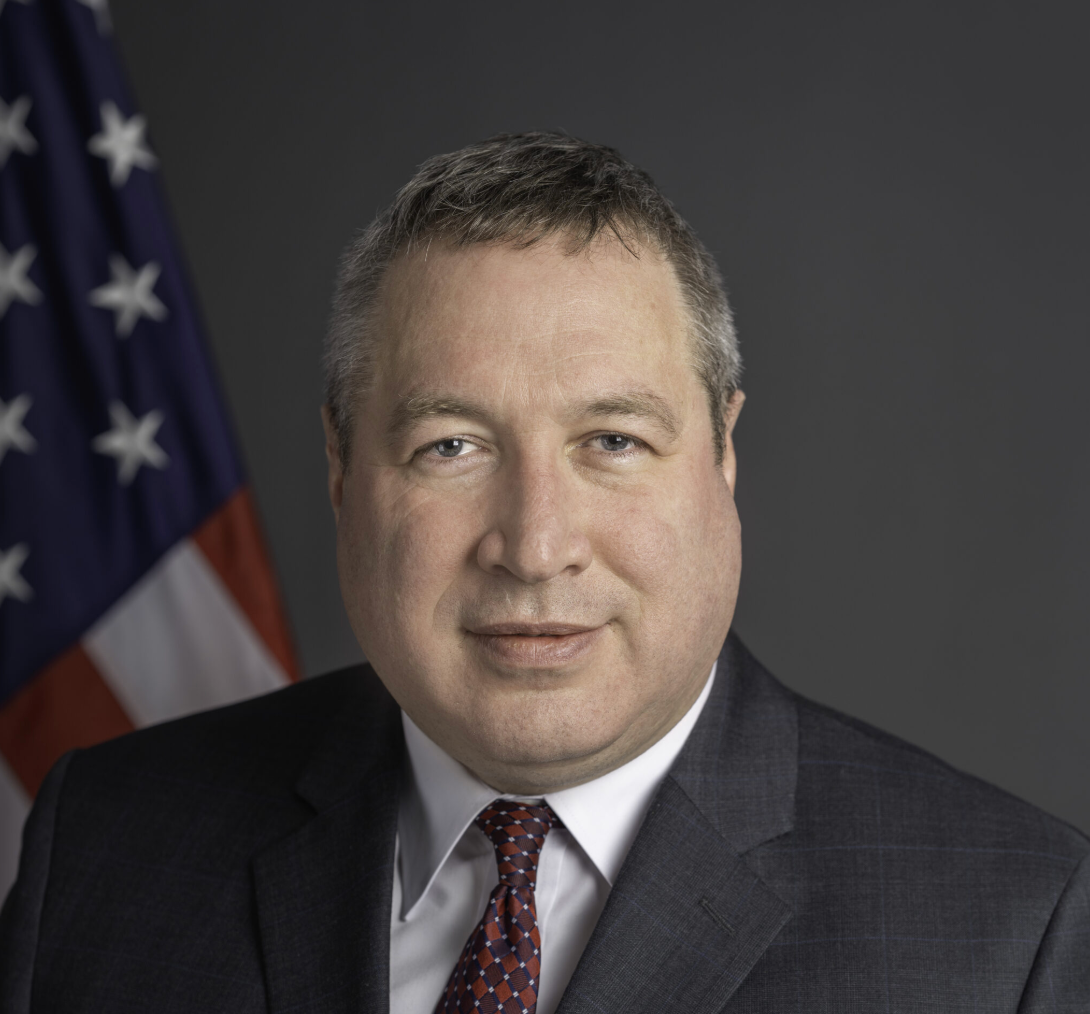 David Gamble usando terno em frente uma bandeira dos Estados Unidos