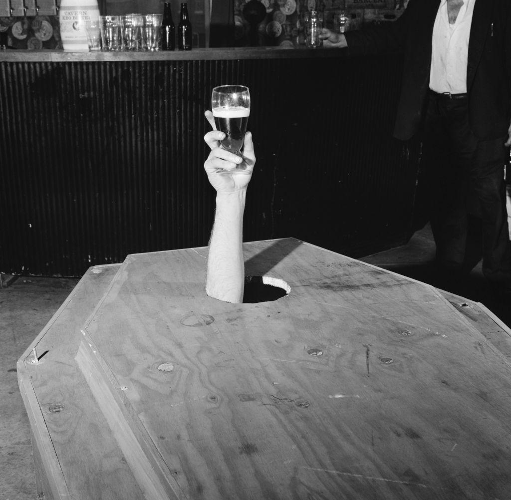 Brazo de Meany saliendo por el hueco del ataúd, sosteniendo una cerveza en su mano.