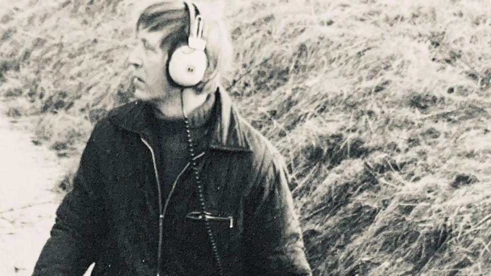 Una imagen en blanco y negro de Dave Derby, que data de la década de 1970 aproximadamente. Mira hacia la izquierda de la imagen y lleva puestos unos auriculares con cable y una chaqueta medio abrochada sobre un jersey de cuello alto. Detrás de él se ve el borde cubierto de hierba de una zanja.