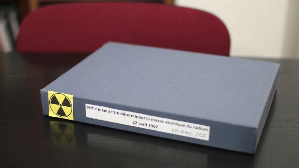 Caixa pequena, na cor cinza, está em cima da mesa. Na lateral há um símbolo amarelo, alertando para a radioatividade