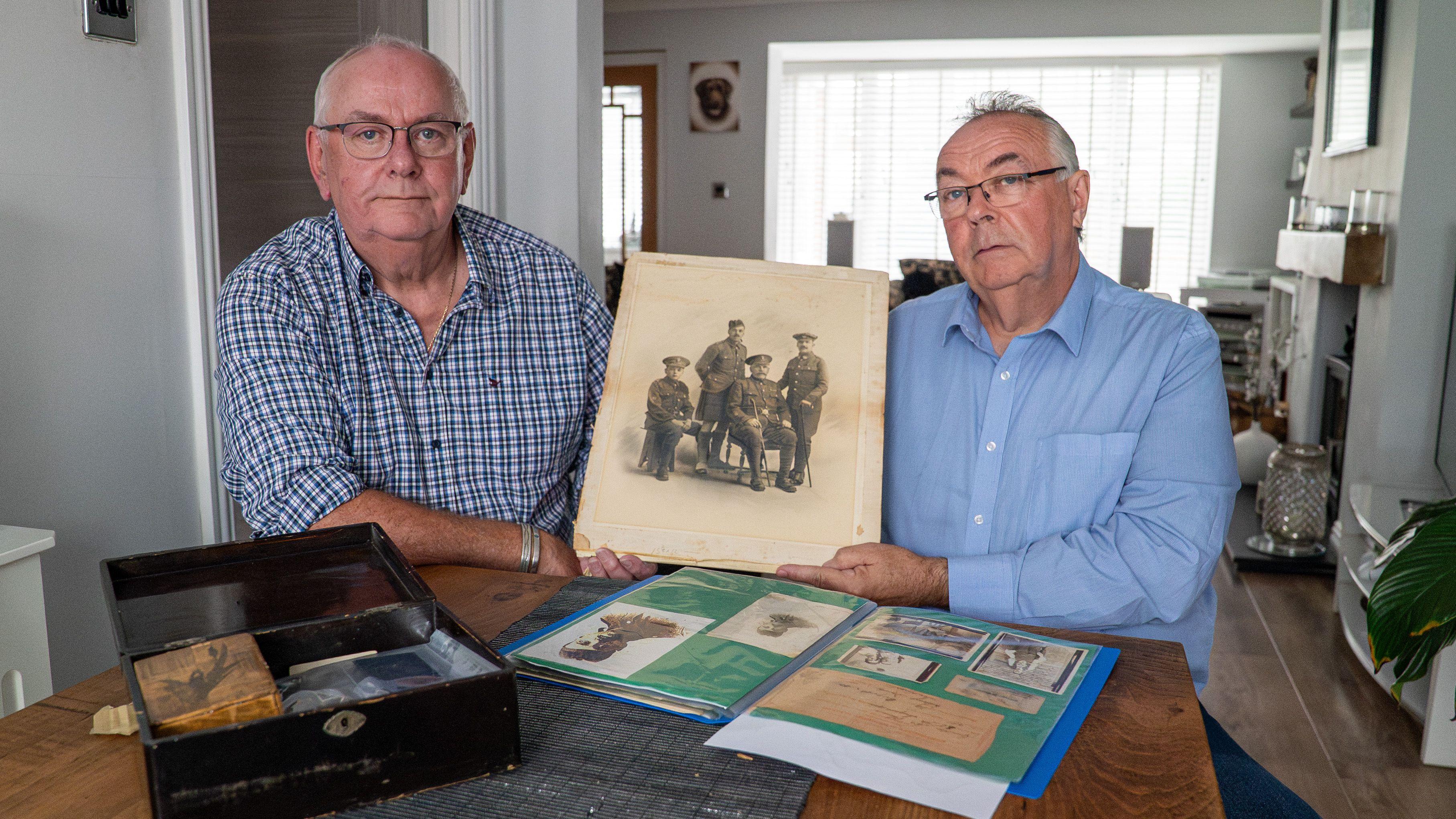 Imagen panorámica de dos hombres de unos 60 años sentados a una mesa de madera en una sala de estar, mientras sostienen una vieja foto sepia de cuatro hombres. Alistair, sentado a la izquierda, lleva una camisa azul a cuadros y Andrew, a la derecha, lleva una camisa azul claro. 