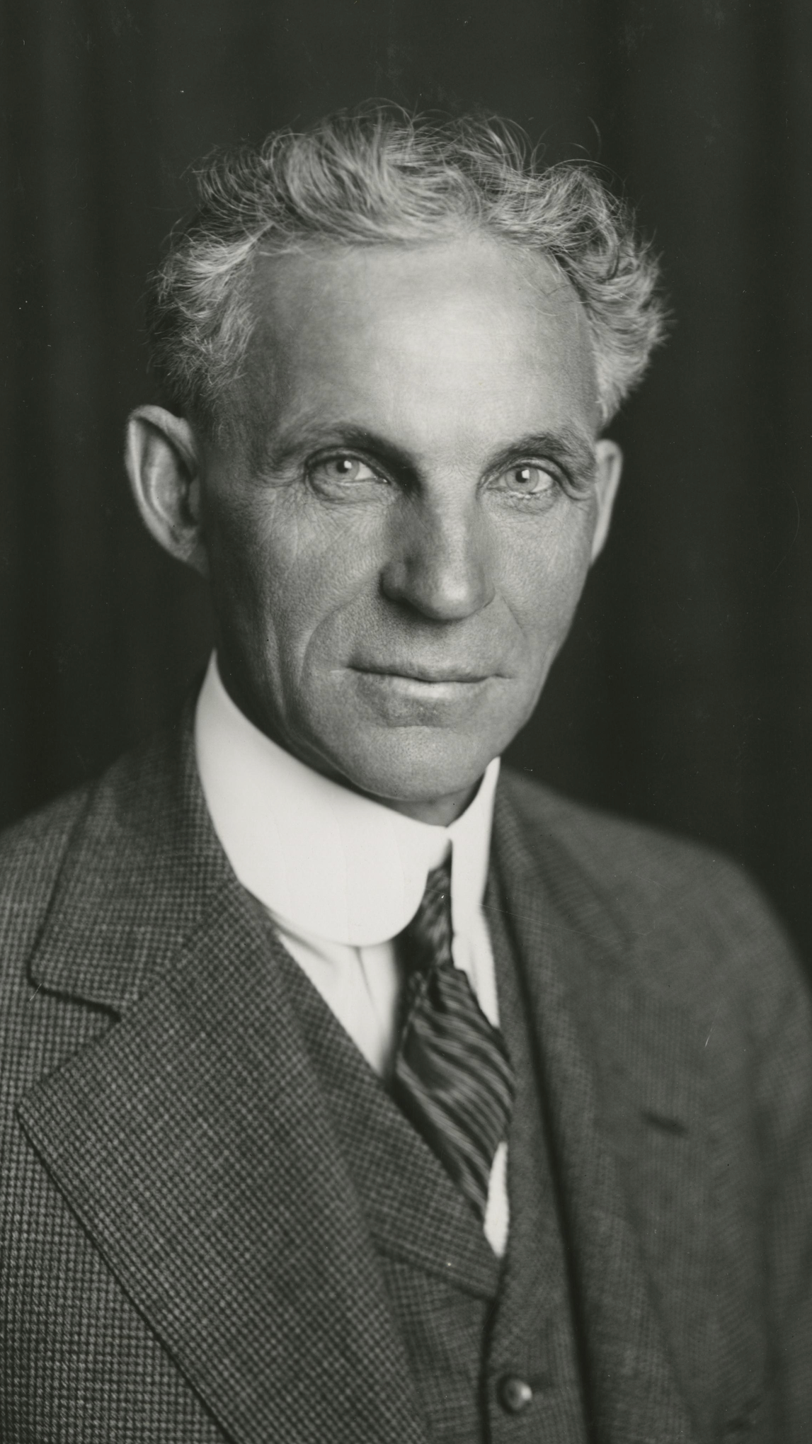 A imagem em preto e branco mostra um homem usando terno, colete, gravata e camisa de colarinho alto posando para um retrato formal. Ele tem cabelos claros e ondulados, penteados para tr&aacute;s, e olha diretamente para a c&acirc;mera com express&atilde;o s&eacute;ria. O fundo escuro e desfocado destaca o rosto e a roupa do retratado.
