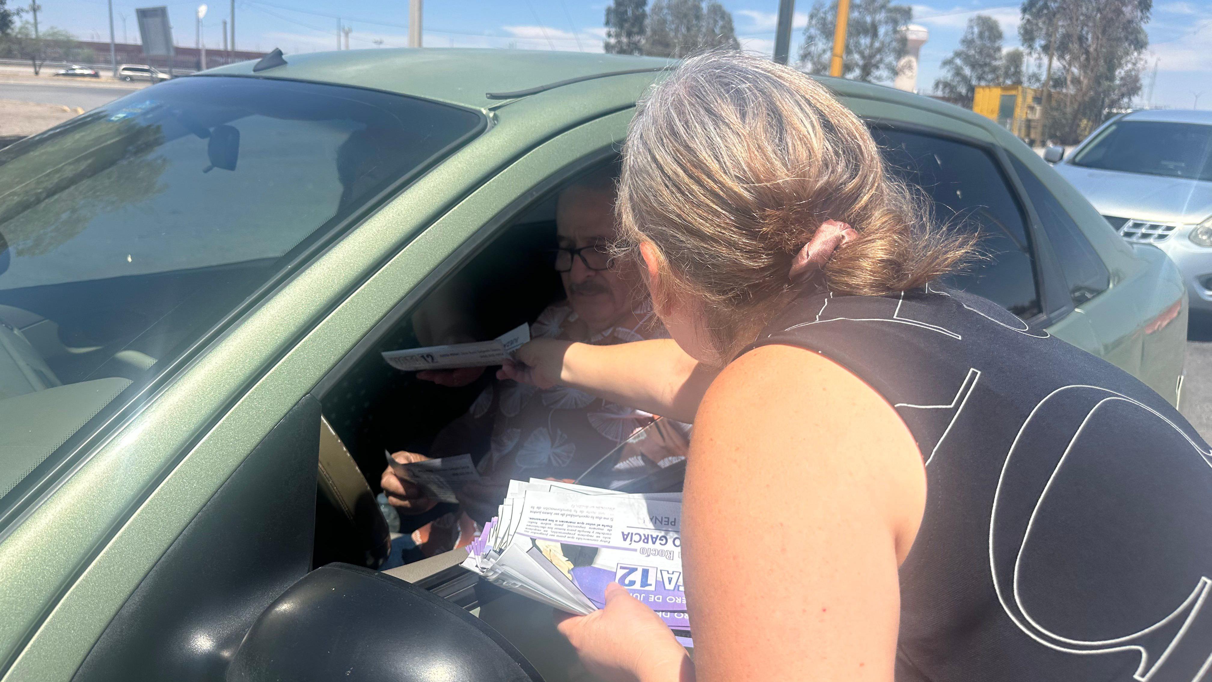 Silvia Delgado, vestindo uma blusa preta e branca, está distribuindo panfletos aos eleitores em um carro na Ponte das Américas.