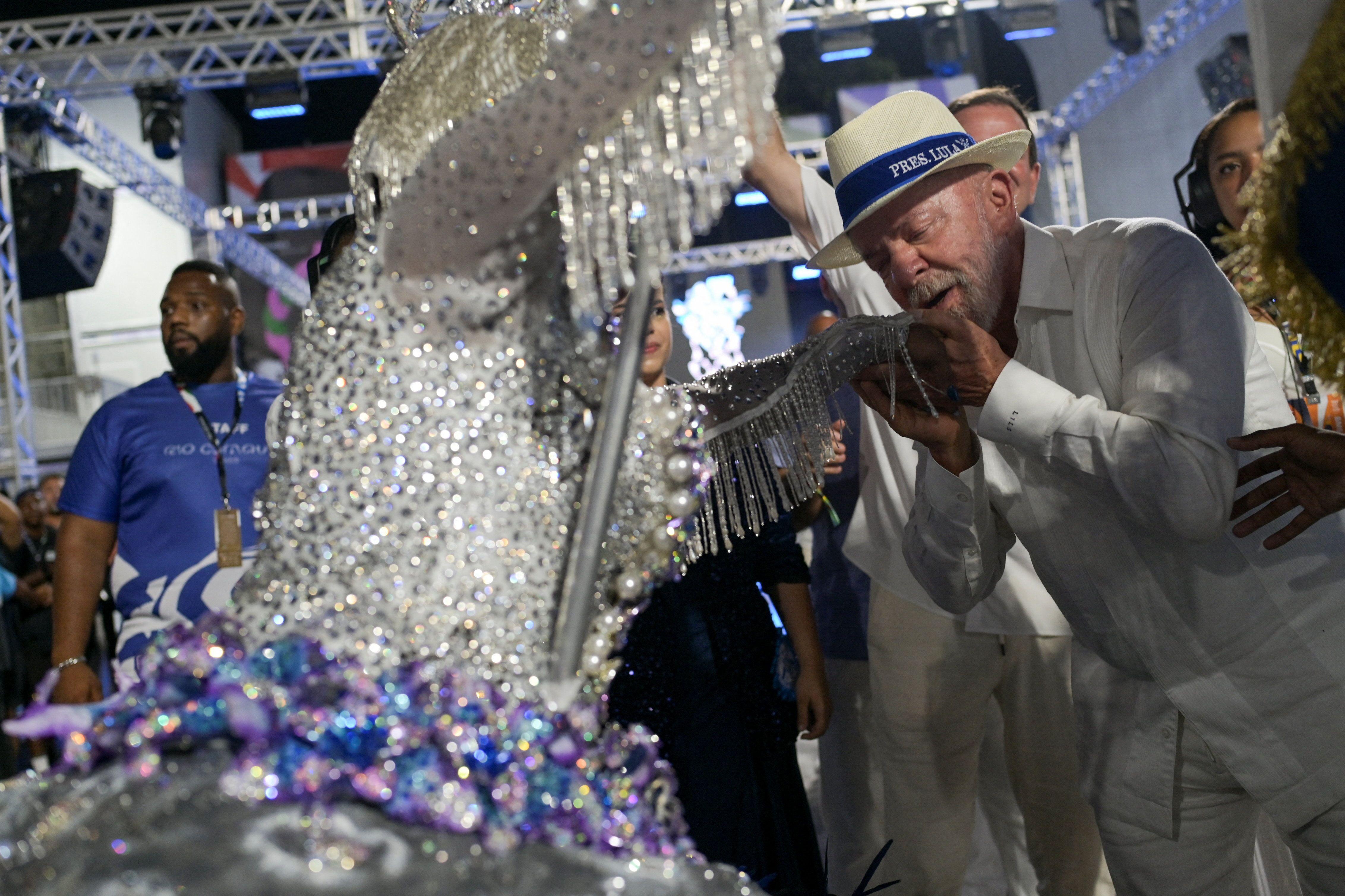 De branco e chap&eacute;u panam&aacute;, Lula cumprimenta porta-bandeira da Acad&ecirc;micos de Niter&oacute;i durante desfile, beijando sua m&atilde;o. Ela usa roupa azul e branca com muito brilho. 
