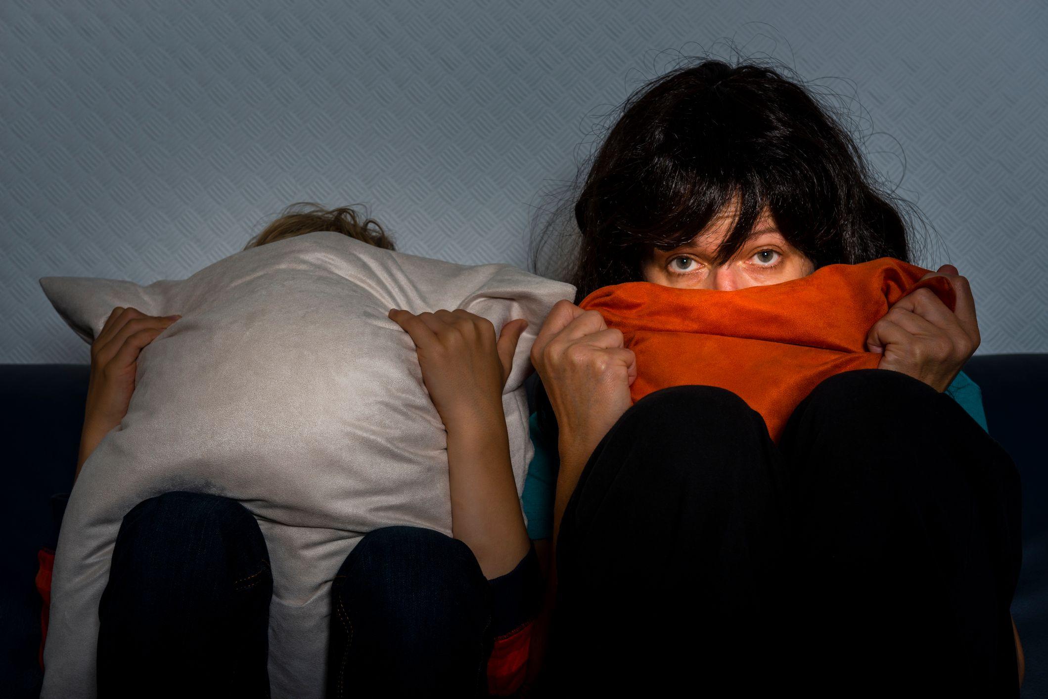 Dos mujeres viendo un programa de telvisión sentadas en una cama. Una de ellas se tapa completamente con una almohada, la otra se lleva una manta hasta la boca pero mira atentamente con ojos asustados